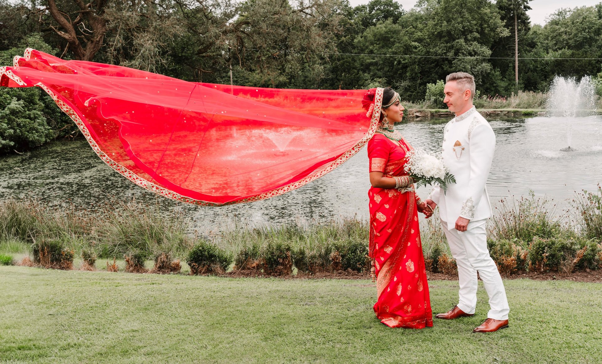 Royal Wedding Cinema Tanzania — couple in traditional wedding attire holding flowing red fabric by waterfront lawn with fountain, Dar es Salaam wedding photography and cinematography