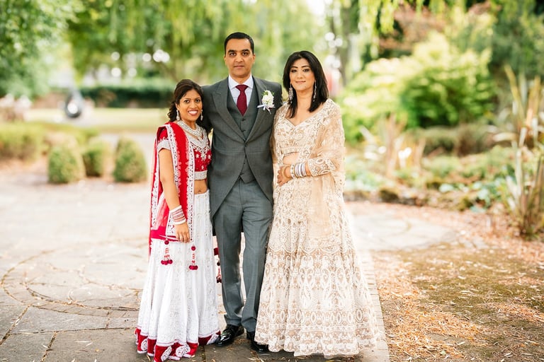 Man in gray suit with boutonniere standing with woman in white embroidered dress and young girl in pink traditional outfit in a garden pathway.