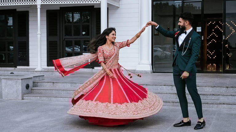 Bride and groom dancing on white building steps, woman in red and gold lehenga spinning, man in dark suit