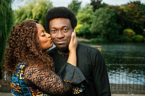 A woman kissing a mans cheek by a river with green trees in the background
