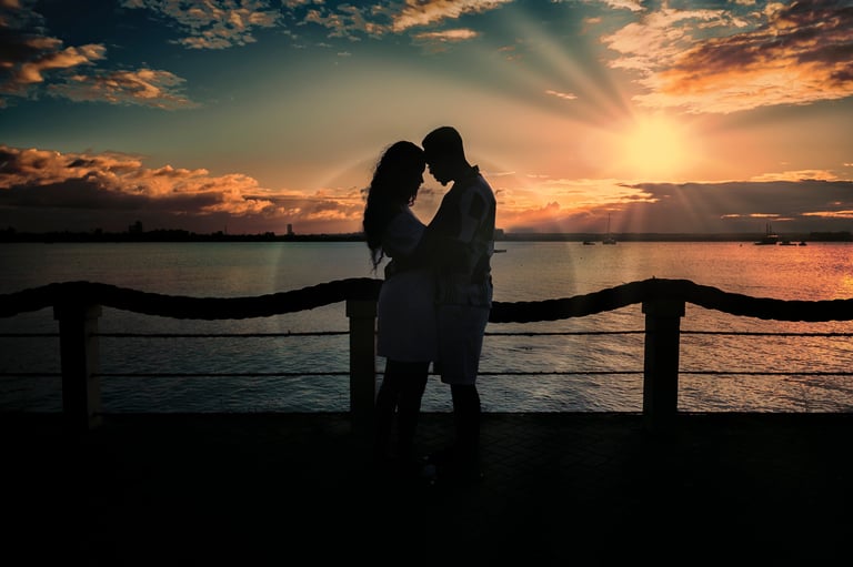 Silhouette of couple kissing at wooden railing against dramatic sunset over water