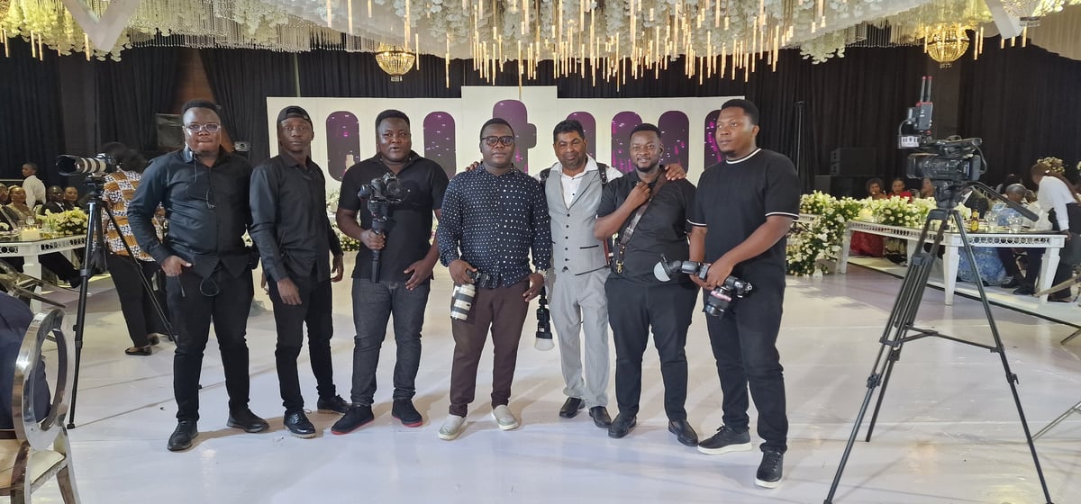 Group of eight men standing together at an elegant event with crystal chandeliers, white flooring, and formal decorations in background
