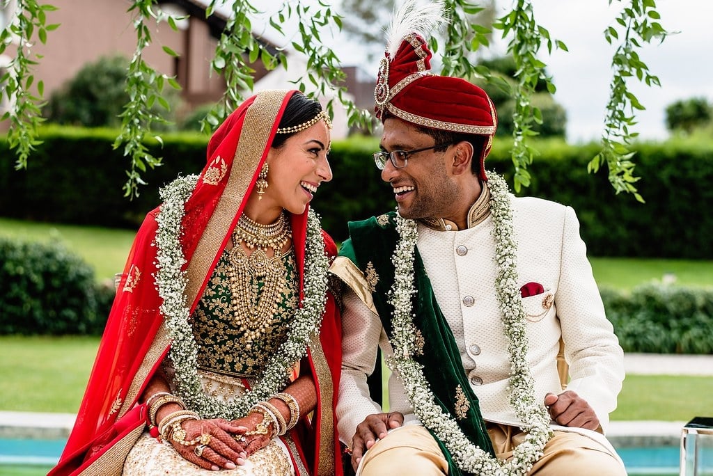 Indian bride and groom in traditional wedding attire sitting together outdoors by a pool, smiling at each other