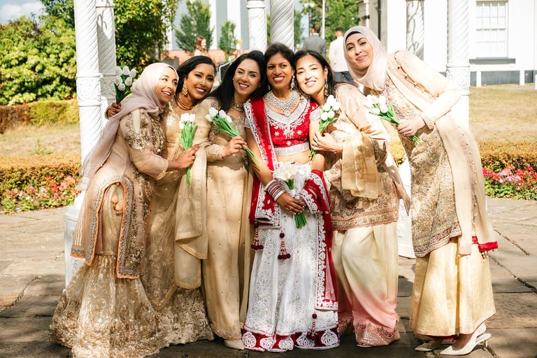 Group of six women wearing traditional South Asian wedding attire posing together outdoors near a columned structure