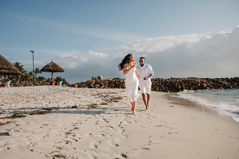 Couple in white clothing running on a sandy beach with tiki umbrellas and rocky pier visible in the background