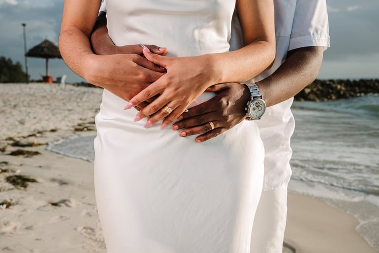 Couple holding hands on beach wearing white clothing, ocean and sand visible in background