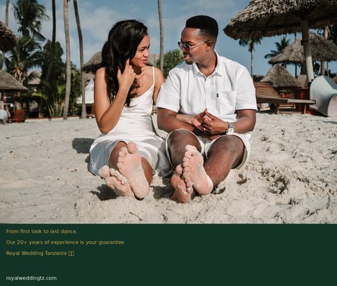 Couple sitting on beach sand facing each other in casual white clothing with palm trees and thatched structures in background