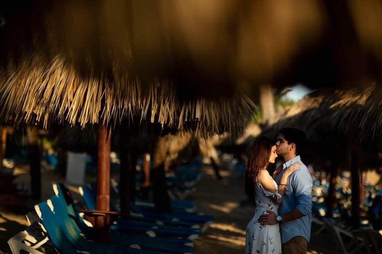 Couple embracing under thatched roof beach shelter with lounge chairs in soft golden light