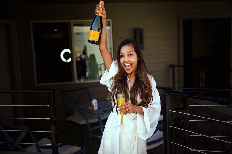 Woman in white shirt holding champagne bottle and glass, celebrating with raised arm on modern patio