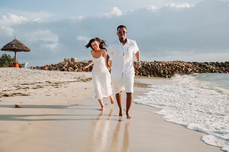 Couple in white clothing running on beach near ocean waves with thatched umbrella and rocky pier in background