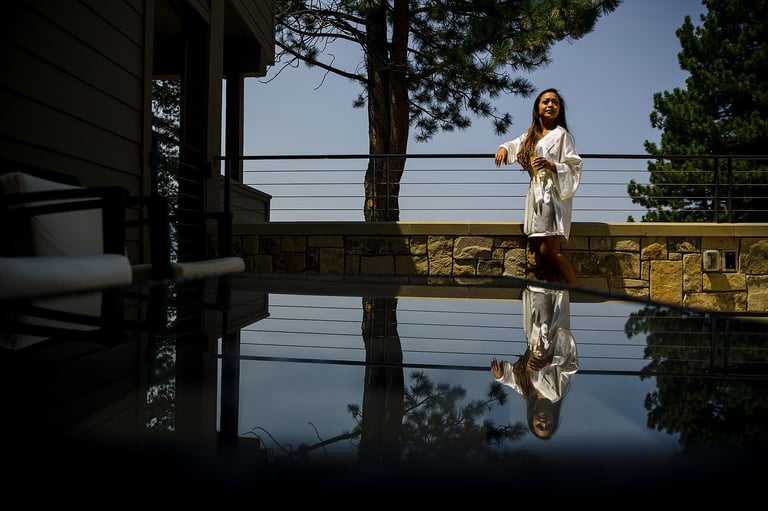 Woman in white outfit standing on a wooden dock, reflected in still water with trees and railing visible