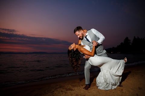 Groom lifting bride on beach during sunset with purple sky and ocean in background