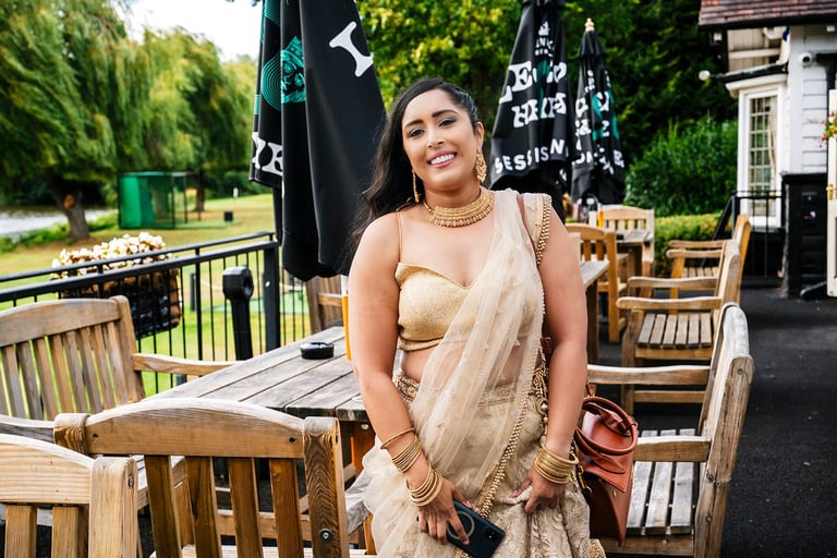 Woman in beige dress smiling on outdoor patio with wooden furniture and river view in background