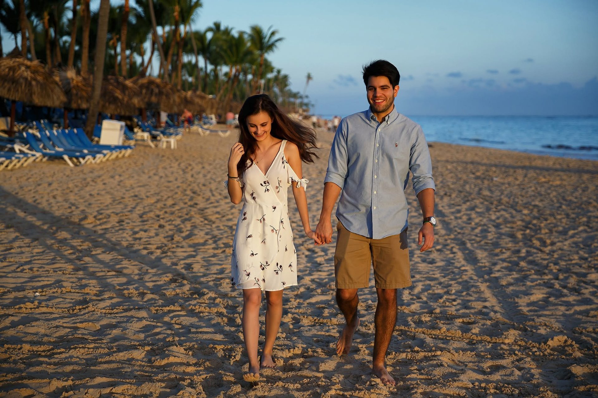 A couple holding hands walking on a sandy beach at sunset with palm trees and blue ocean in the background