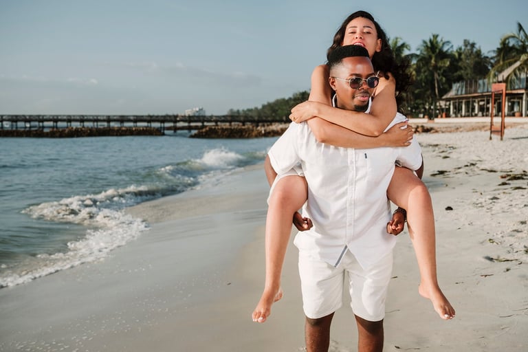 Man in white shirt giving piggyback ride to woman on sandy beach with ocean waves and pier in background