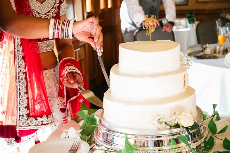 Three-tiered white wedding cake with gold accents and white flowers, displayed at an Indian wedding reception with bride and groom in traditional attire