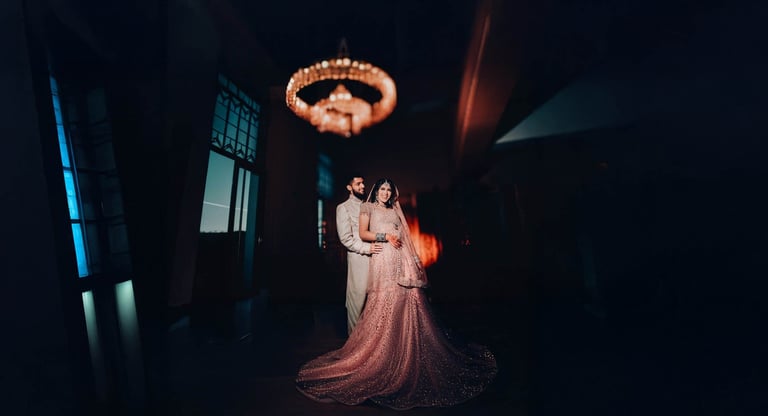 Bride and groom dancing under warm overhead lighting in a dark venue, bride in flowing pink gown and groom in beige suit