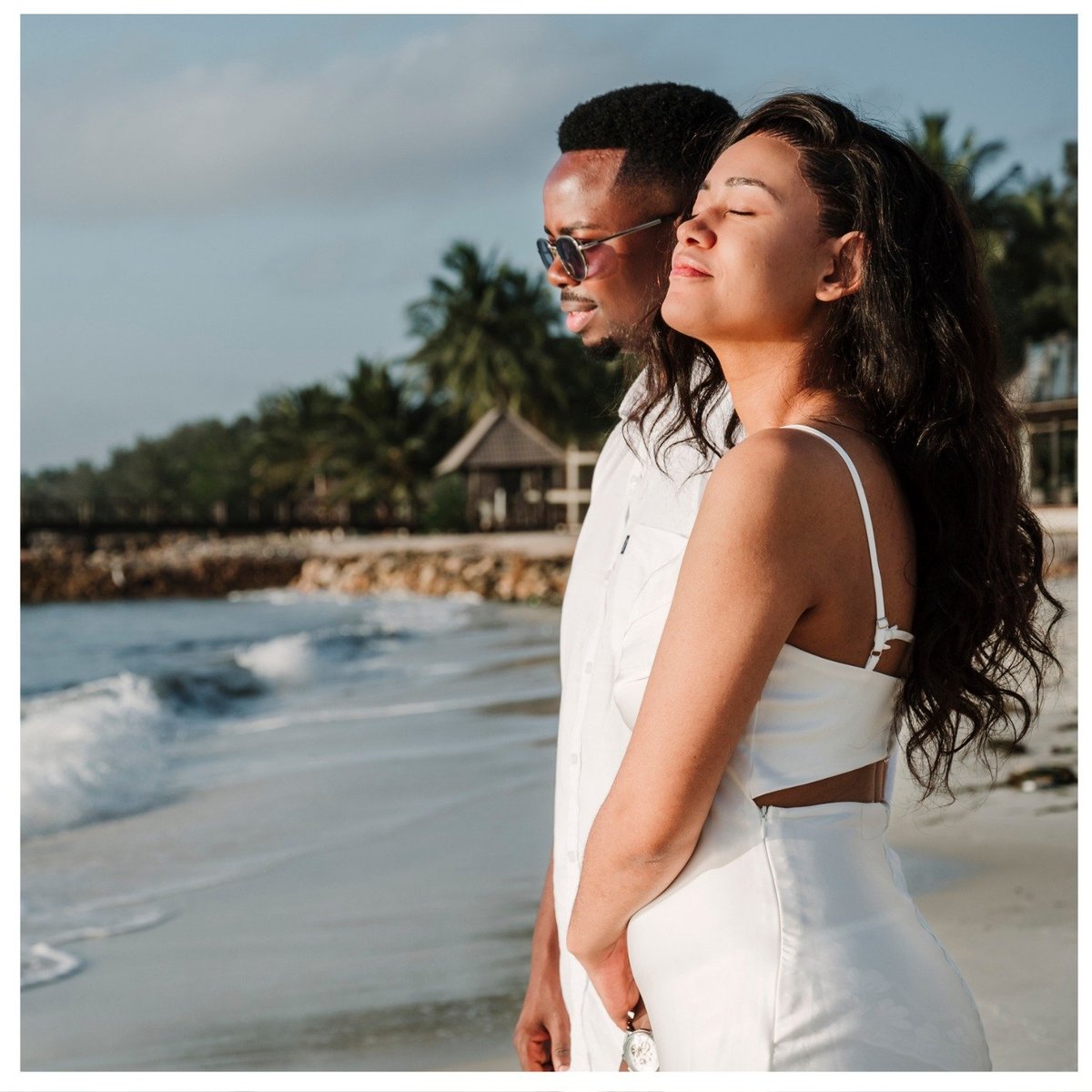 Couple in white outfits standing together on a tropical beach with palm trees and calm ocean waves in the background