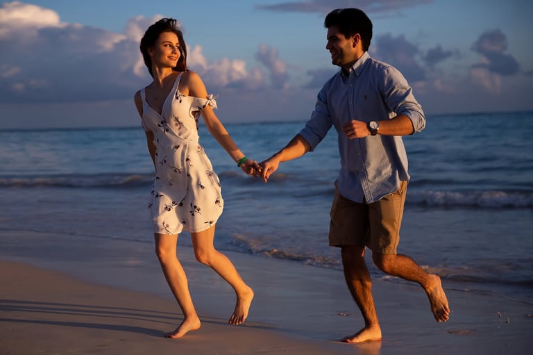 Couple holding hands while running on beach at sunset with ocean and sky in background