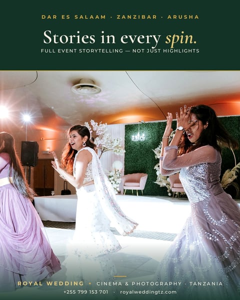 Bride in white embellished gown dancing with guests in celebration at an elegant wedding venue with warm orange lighting and green foliage backdrop in Tanzania