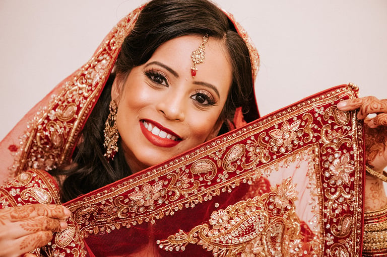 Woman in traditional Indian bridal attire with red and gold embroidered dupatta, bindi, and jewelry smiling at camera