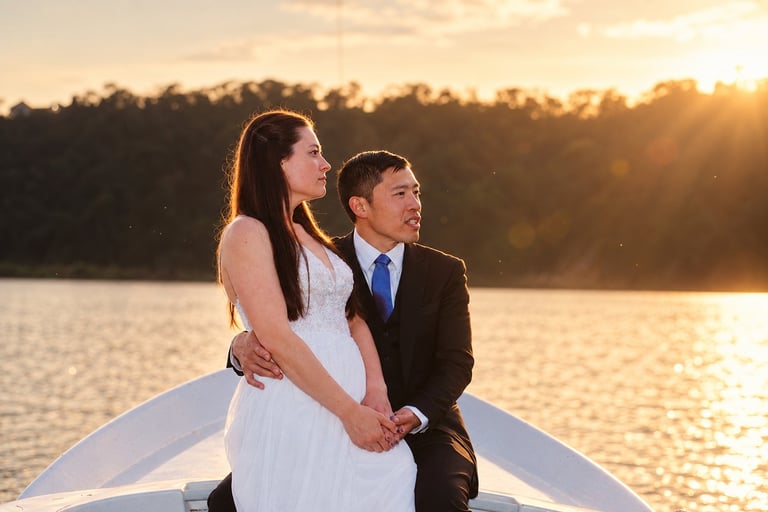 Newlyweds in wedding attire standing on a boat at sunset with water and forested shoreline in background