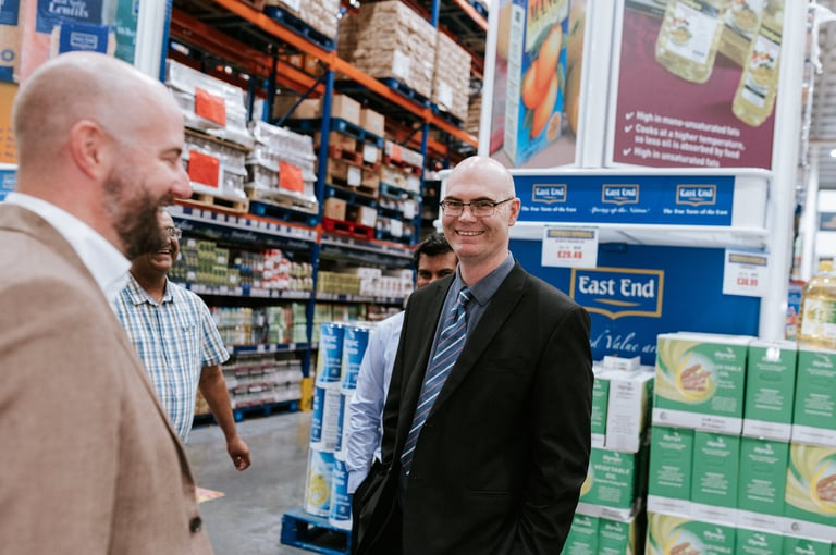 Corporate warehouse photography — two business men conversing in warehouse store with merchandise shelves stacked in background, Dar es Salaam