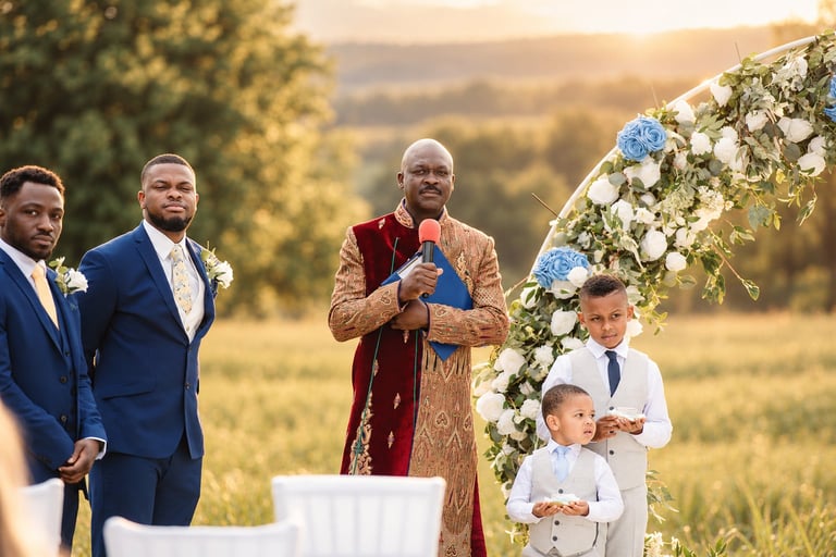 Wedding photographer capturing an outdoor ceremony at golden hour, with the priest and groomsmen standing at a floral arch and two young boys in white suits in the foreground against a sunset countryside landscape in Tanzania