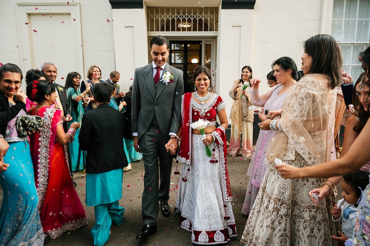 Groom in dark suit walking with bride in white dress through a corridor surrounded by guests in colorful traditional wedding attire