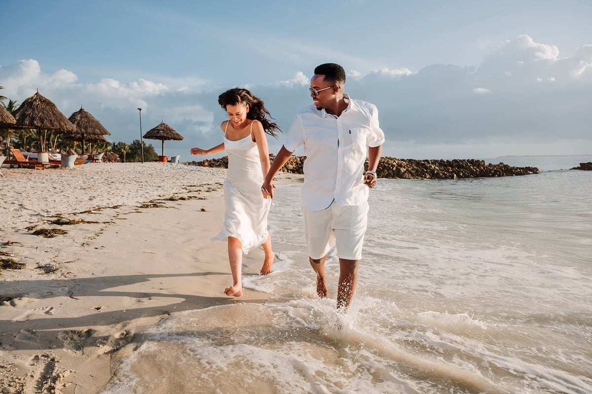 Couple in white clothing running into the ocean on a tropical beach with palm-thatched umbrellas in the background