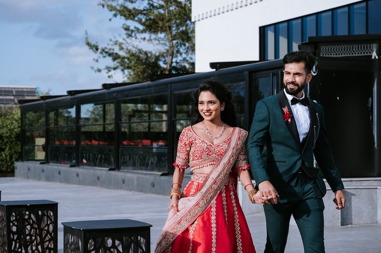 Bride in pink and red lehenga and groom in dark teal suit walking hand in hand outside modern building