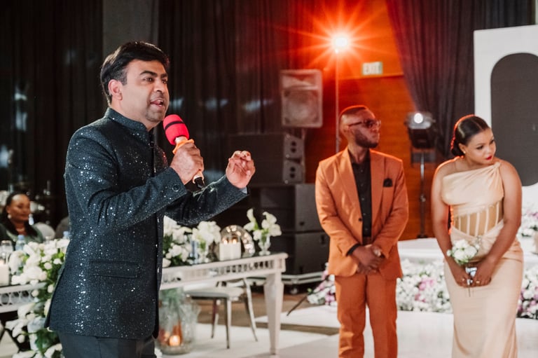 Man in dark sequined jacket speaking at microphone on stage with three other people in formal attire and floral decorations