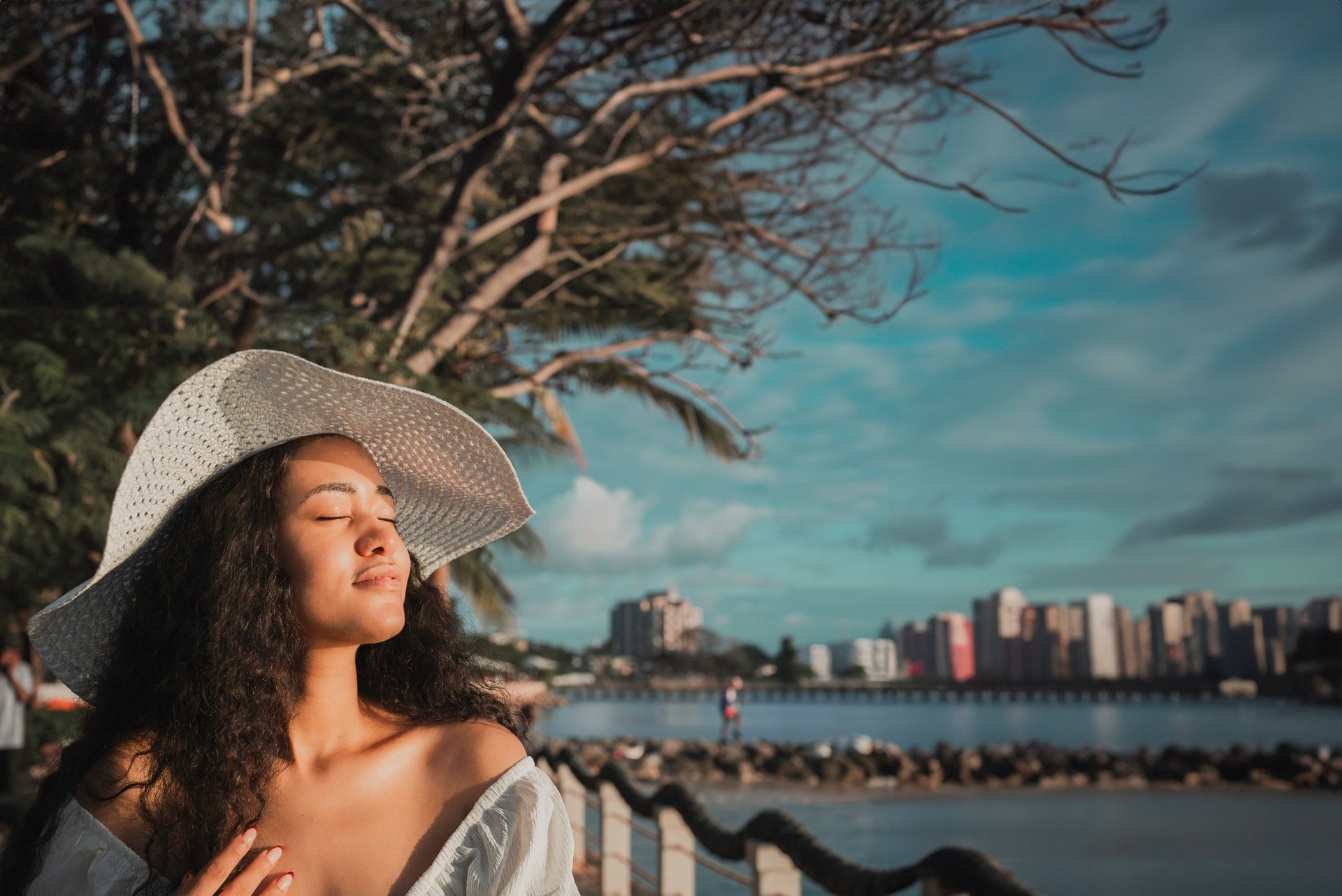 Woman in white hat overlooking waterfront city skyline with bare trees and blue sky