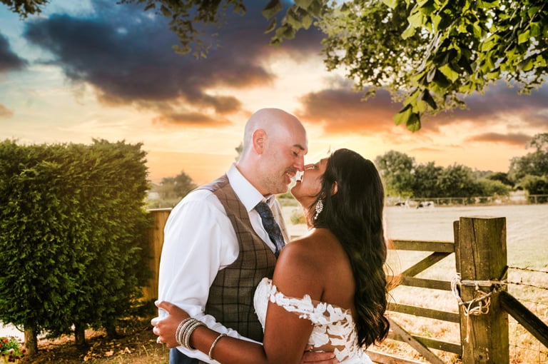 Bride and groom kissing outdoors at sunset near a wooden fence and tree-lined landscape