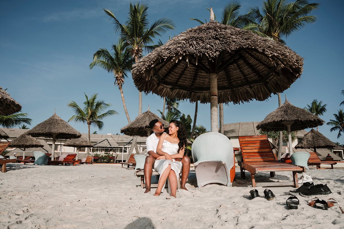 Couple sitting together on beach under large thatched umbrella with palm trees and beach cabanas in background