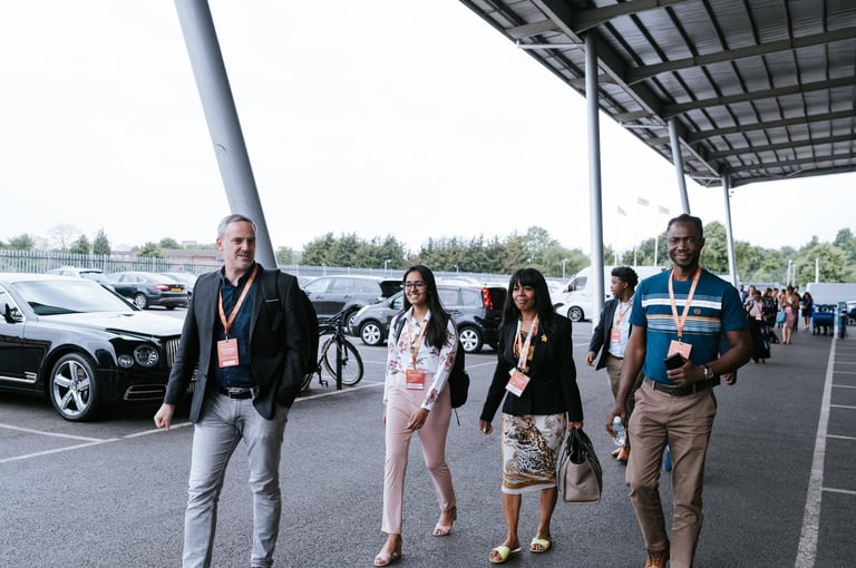 Corporate event logistics photography — group of delegates wearing ID badges walking through parking lot under covered structure with vehicles, Tanzania