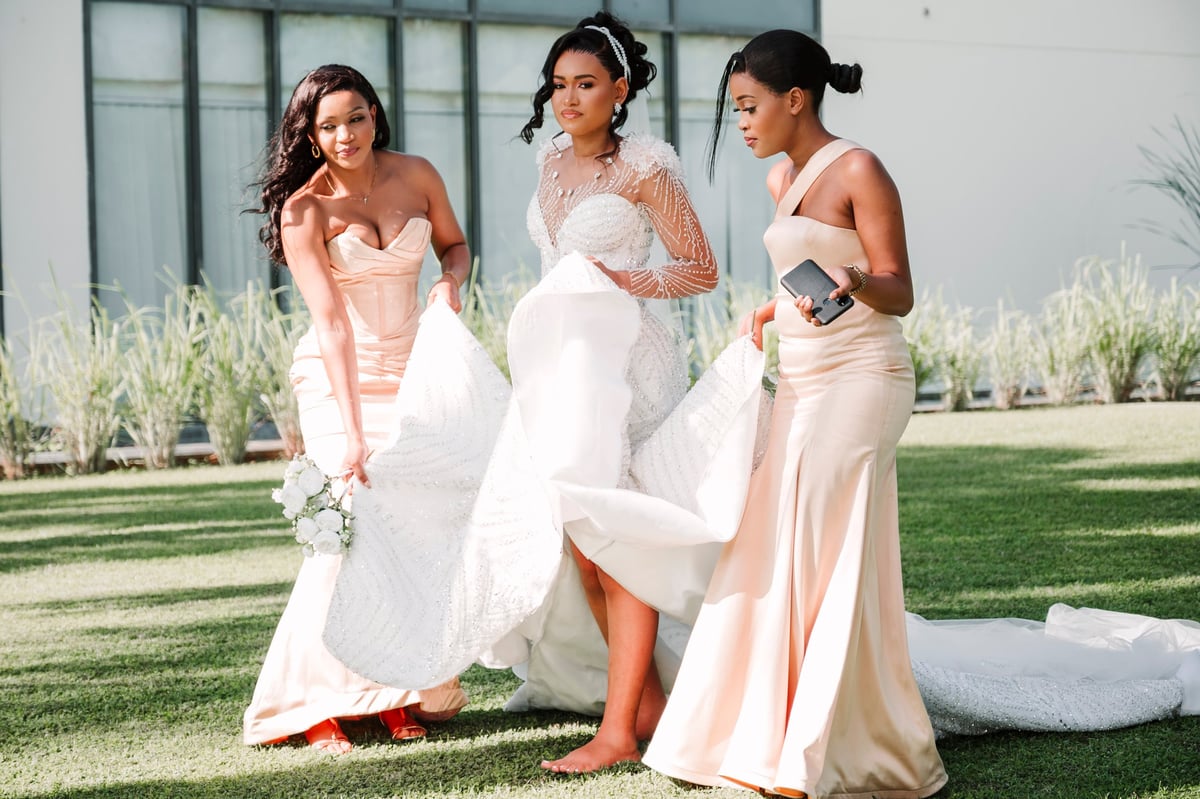 Three women in formal attire posing on a lawn, with the center woman in a white wedding dress