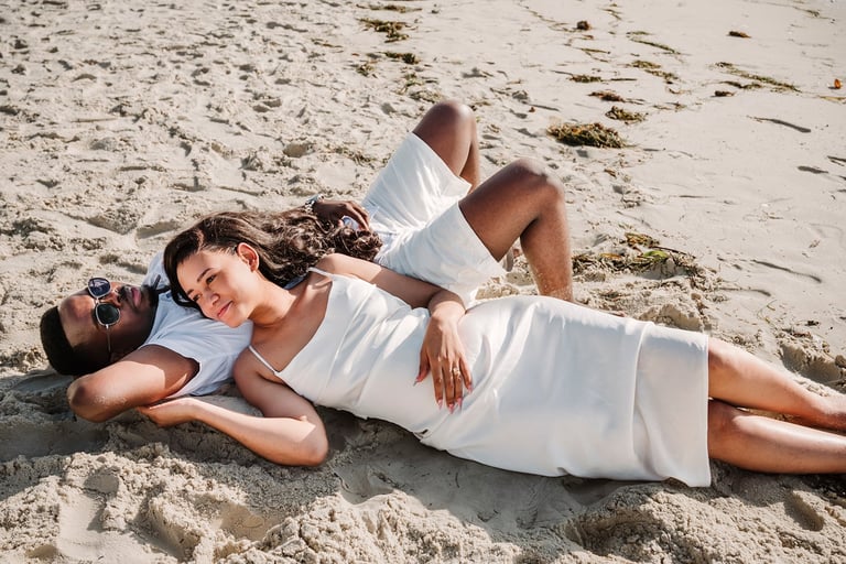 Two women in white clothing lying on sandy beach, smiling at camera during daytime photoshoot
