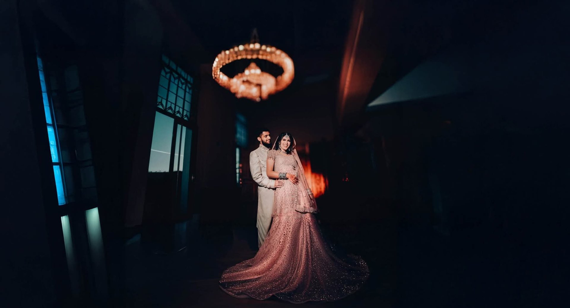 Bride and groom embrace under an ornate circular chandelier in a dimly lit hallway with dramatic lighting