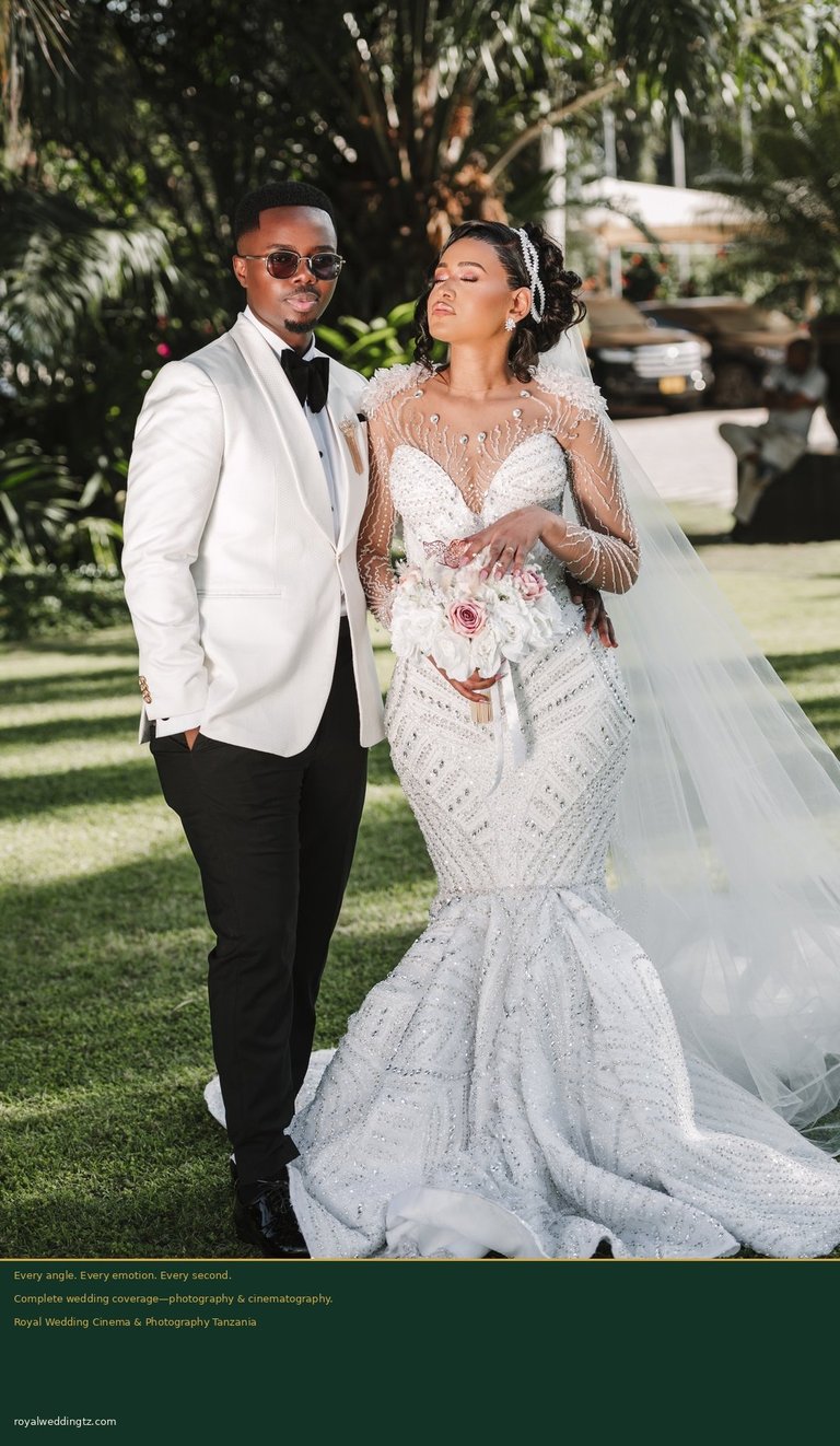 Groom in white jacket and bride in beaded mermaid gown with veil walking together on green lawn at outdoor wedding ceremony.