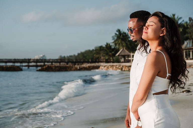 Couple standing together on a tropical beach looking out at the ocean with palm trees and a pier in the background