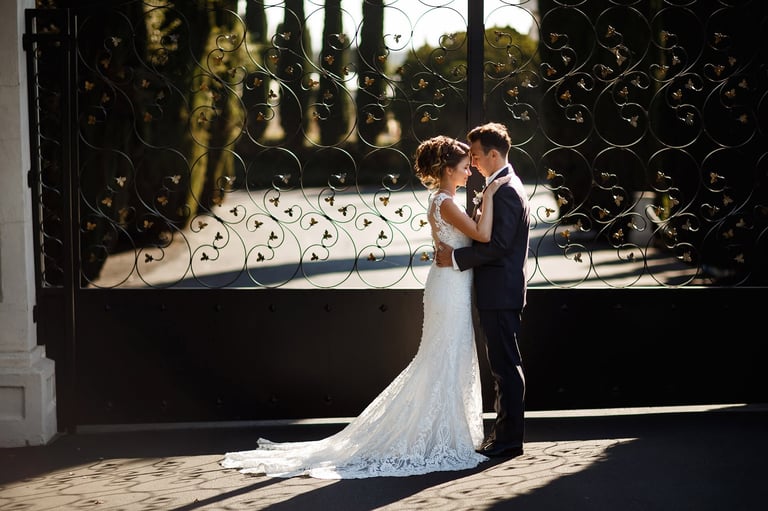 Bride and groom embrace in front of ornate black gate with lace-draped backdrop on sunny day