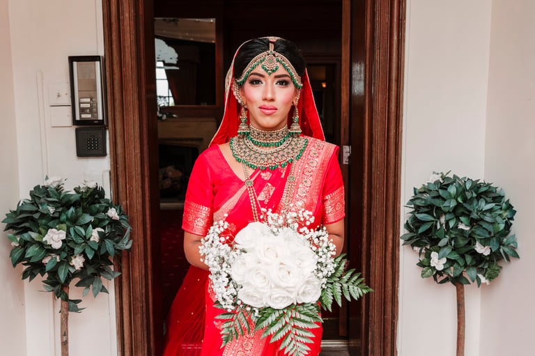 Bride in traditional red wedding attire with gold embroidery holding white bouquet in doorway with floral arrangements — Asian wedding photography Dar es Salaam Tanzania by Royal Wedding Cinema & Photography