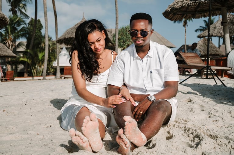 Couple sitting on beach sand holding hands, wearing white clothing, tropical resort setting with palm trees and thatched umbrellas