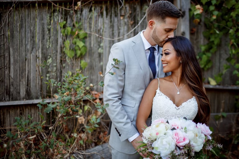 Bride and groom posing together outdoors in front of a wooden fence, holding a pink and white bouquet