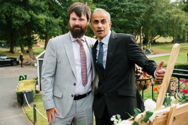 Two men in business suits posing together outdoors in a park setting with flowers and greenery in the background.