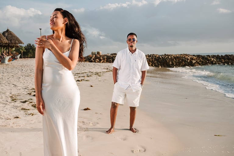 A couple in white clothing standing on a sandy beach with waves in the background during daytime