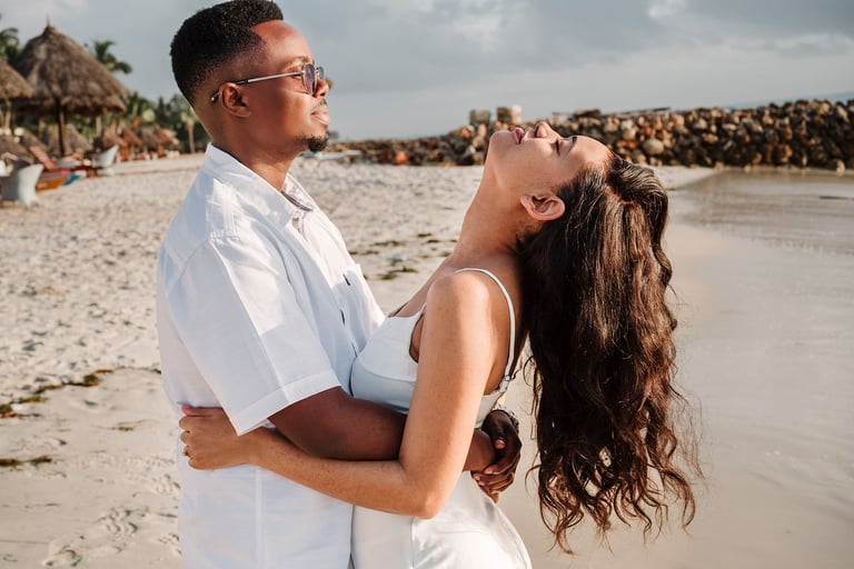 Couple in white clothing embracing on a sandy beach during daytime, with palm trees and people visible in the background