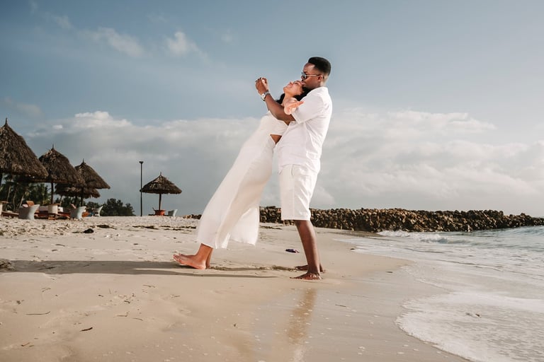 Groom in white shirt and pants lifts bride in white dress on a tropical beach at the waters edge