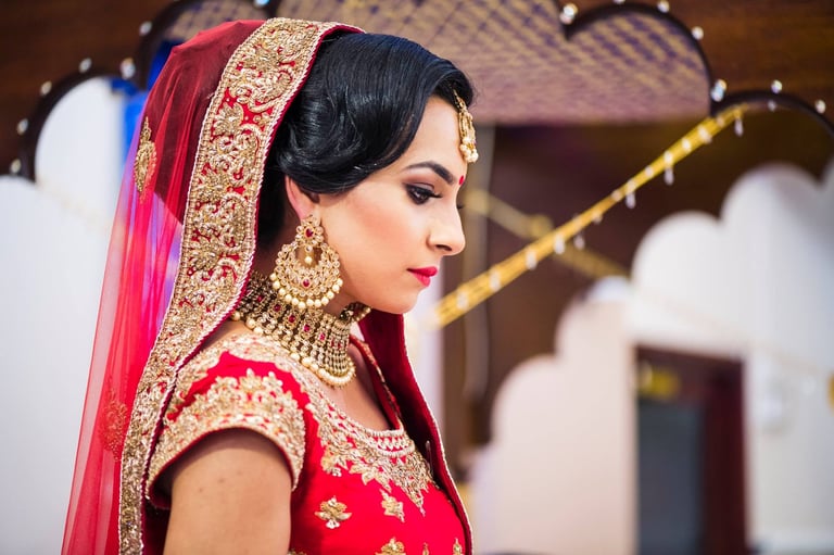 Woman in traditional red bridal lehenga with gold embroidery, jewelry, and pink veil, photographed in profile at an indoor venue